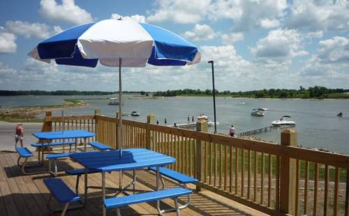 A blue and white umbrella shades a picnic table overlooking a lake with boats and people enjoying the sunny day.
