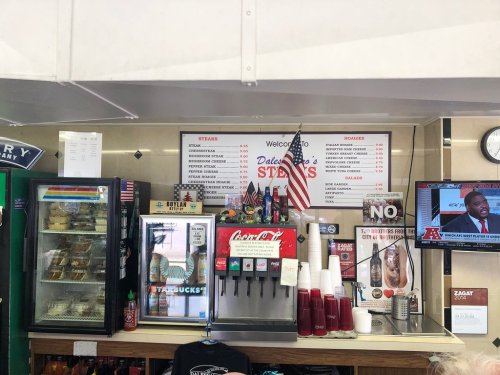 A food counter displaying a menu for steaks and burgers, with a drink station and a TV showing a sports program.
