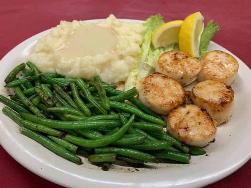A plate of seared scallops with green beans, mashed potatoes topped with gravy, and lemon wedges on a red tablecloth.