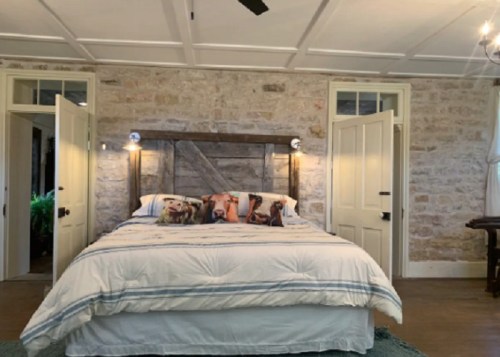 Cozy bedroom with a stone wall, a wooden bed frame, and decorative pillows featuring dogs. Bright and inviting atmosphere.
