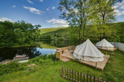 Two large tents on wooden decks by a serene river, surrounded by lush green trees and a clear blue sky.