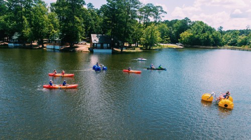 A serene lake scene with people kayaking and paddleboarding, surrounded by trees and a cabin in the background.