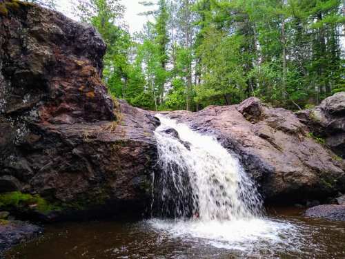 A small waterfall cascades over rocky terrain, surrounded by lush green trees and a tranquil pool below.