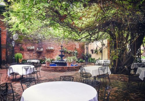 A charming outdoor dining area with tables, a fountain, and lush greenery in a brick-walled courtyard.