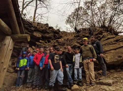 A group of people, including children and adults, poses together in front of a rocky hillside in a wooded area.