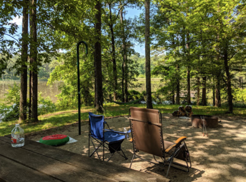 A serene campsite by a lake, featuring two chairs, a water bottle, and a fire pit surrounded by trees.