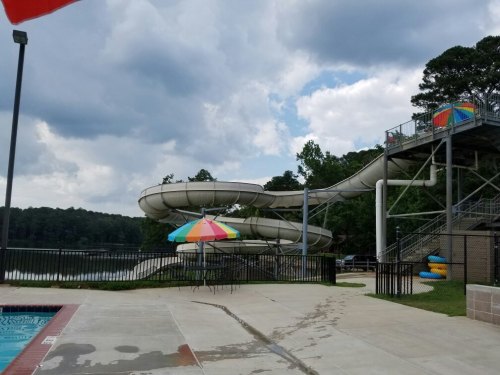 Water slide at a poolside with colorful umbrellas, surrounded by trees and a cloudy sky.