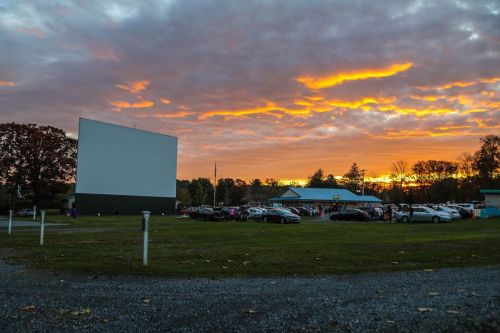 A drive-in movie theater at sunset, with cars parked and a colorful sky filled with clouds.