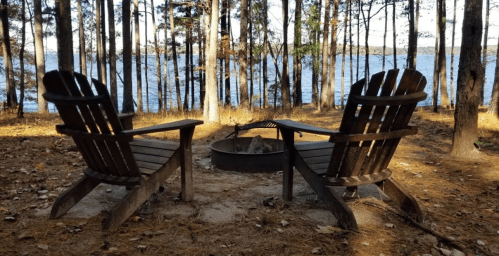 Two wooden chairs facing a fire pit by a lake, surrounded by trees and fallen leaves.