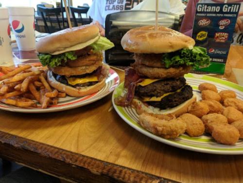 A towering burger with lettuce, bacon, and onion rings, served with fries and chicken nuggets on a wooden table.