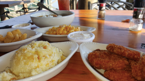 A table with various dishes: fried chicken, macaroni and cheese, mashed potatoes, and snacks, with drinks in the background.
