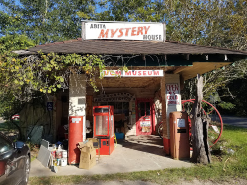 Abita Mystery House exterior with vintage signs, red doors, and surrounding greenery on a sunny day.