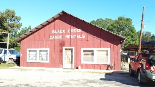 Red wooden building with "Black Creek Canoe Rentals" sign, surrounded by trees and parked vehicles. Clear blue sky.