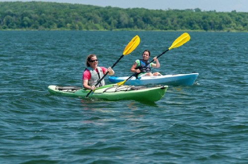 Two people kayaking on a calm lake, one in a green kayak and the other in a blue kayak, enjoying a sunny day.