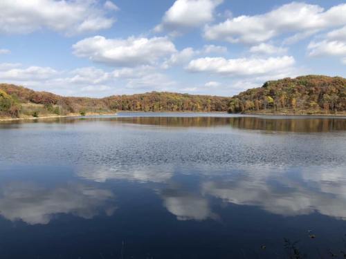 A serene lake reflecting fluffy clouds and autumn-colored trees under a blue sky.