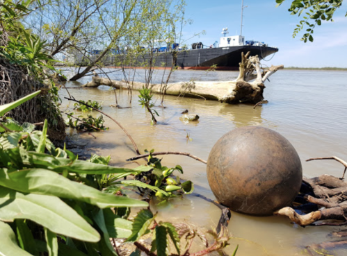 A large metal sphere rests on the riverbank, with a boat in the background and lush greenery along the water's edge.