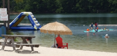 A sandy beach scene with a red chair, a straw umbrella, and people on paddleboards near an inflatable water slide.