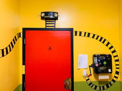 A bright yellow wall features a red door with a playful, cartoonish design and a black and yellow striped arm extending from it.
