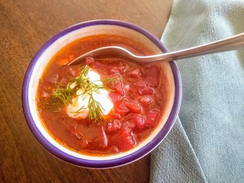 A bowl of red soup topped with sour cream and fresh dill, with a spoon resting inside, on a wooden table.