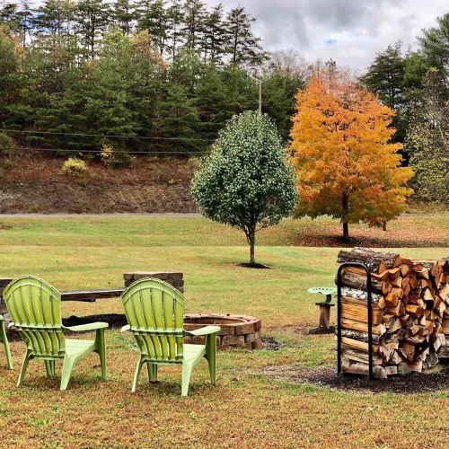 Two green chairs face a fire pit, with a stack of firewood nearby and colorful autumn trees in the background.