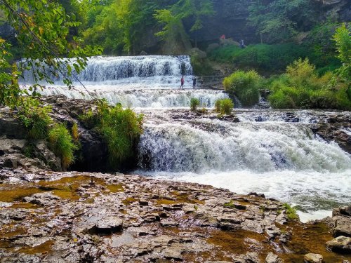 A serene waterfall cascades over rocky tiers, surrounded by lush greenery and a person standing in the water.