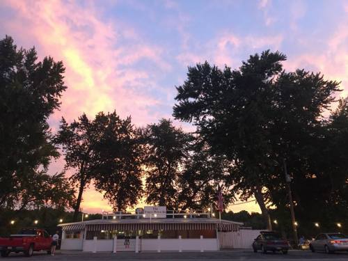 A charming building surrounded by tall trees, with a colorful sunset sky in the background. Cars are parked nearby.
