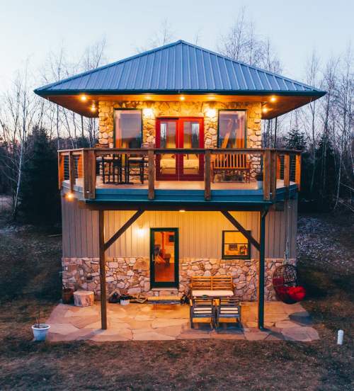 A cozy two-story cabin with a stone exterior, balcony, and warm lights, surrounded by trees at dusk.
