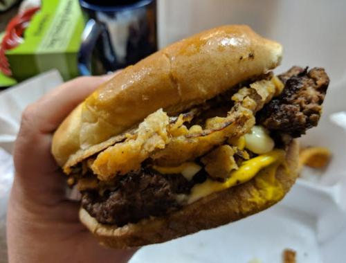 A hand holding a large burger with a beef patty, cheese, crispy onion rings, and mayonnaise on a toasted bun.