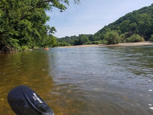 A serene river scene with lush greenery on both sides and a kayaker in the distance under a clear blue sky.