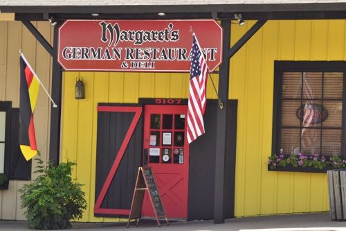 Exterior of Margaret's German Restaurant & Deli, featuring a red door, flags, and colorful flower boxes.
