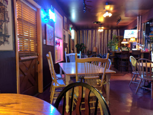Cozy restaurant interior with wooden tables, chairs, and warm lighting, featuring a bar area in the background.
