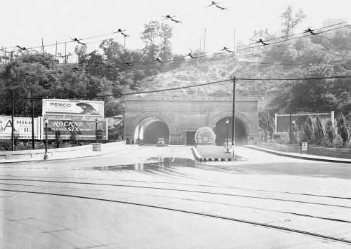 Black and white photo of a tunnel entrance with billboards, power lines, and trees on a hillside in the background.