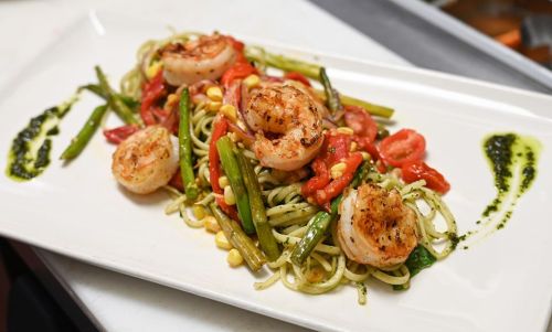 A plate of pasta topped with grilled shrimp, asparagus, cherry tomatoes, and drizzled with pesto sauce.