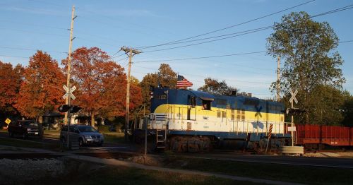 A blue and yellow train engine passes a crossing with autumn trees and a car nearby, under a clear sky.
