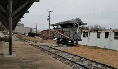 A train station platform with a vintage-style shelter and nearby buildings, set along a gravel railway track.