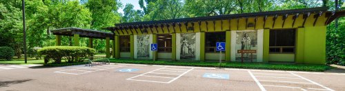 A green building with large windows, featuring murals, surrounded by trees and accessible parking spaces.