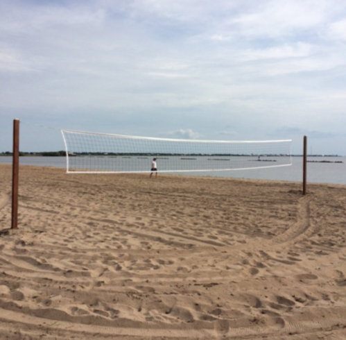 A beach volleyball net stands on sandy shores, with a person in the distance near the water under a cloudy sky.