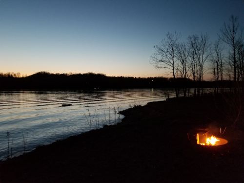 A serene lakeside scene at dusk, with a glowing campfire and silhouettes of trees against a calm water surface.