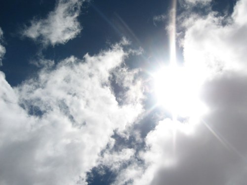 Bright sun shining through fluffy white clouds against a deep blue sky.