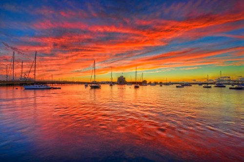 A vibrant sunset over a calm harbor, with colorful clouds reflecting on the water and boats anchored nearby.