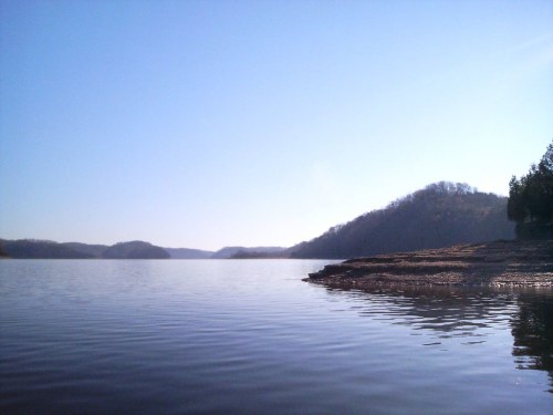 A serene lake scene with calm water reflecting the clear blue sky and distant hills on the horizon.