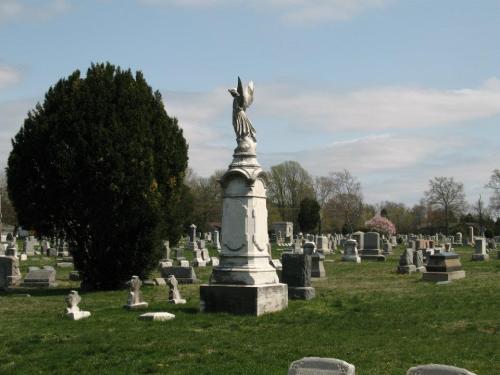 A cemetery scene featuring a tall angel statue surrounded by gravestones and lush greenery under a cloudy sky.