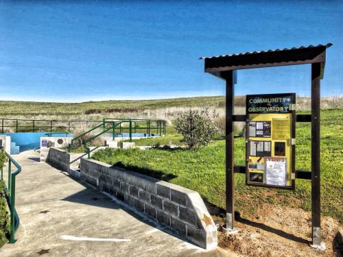 A community observatory entrance with a sign, pathway, and green hills under a clear blue sky.