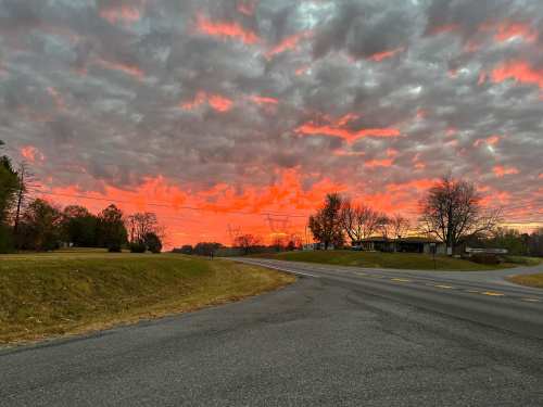 A vibrant sunset with fiery orange and red hues against a cloudy sky, viewed from a roadside.