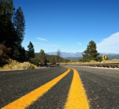 A winding road with yellow lines, flanked by trees and mountains under a clear blue sky.