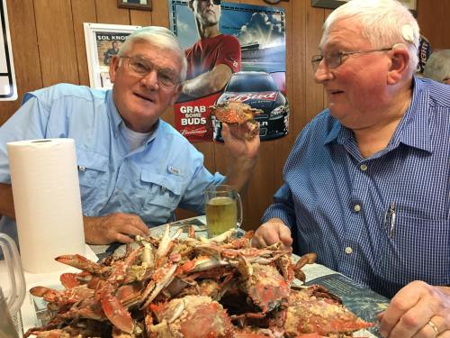 Two older men sit at a table filled with crabs, smiling and enjoying a meal together in a cozy restaurant setting.