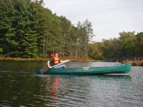A person in an orange life jacket paddles a green canoe on a calm lake surrounded by trees.