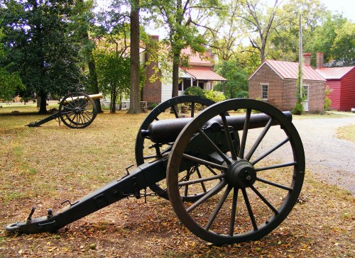 Two historic cannons on display in a grassy area, with brick houses and trees in the background.
