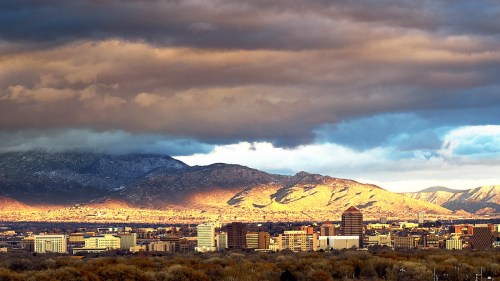 A city skyline with mountains in the background, illuminated by sunlight under dramatic clouds.