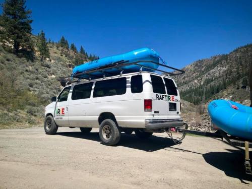 A white van with a blue raft on top parked in a scenic mountainous area.